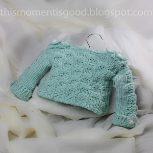 Può includere: Un maglione di lana per neonato di colore verde chiaro con fiori bianchi all'uncinetto sulle spalle e sui polsini.