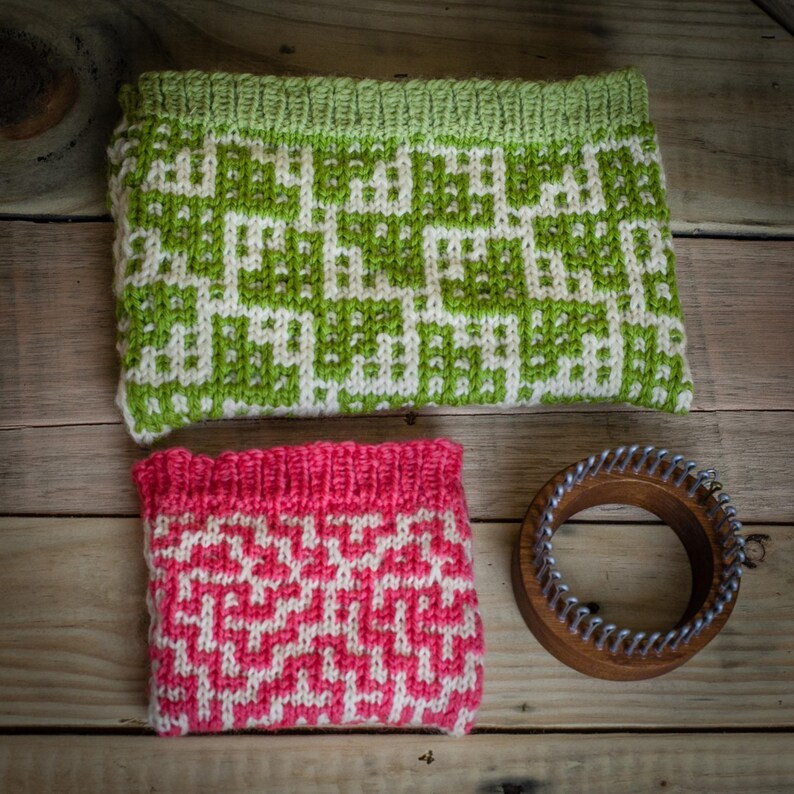 May include: Two knitted items with geometric patterns in green and pink, alongside a wooden knitting loom. The green item is rectangular, with a white and green design. The pink item is smaller, with a white and pink design. The loom is round.