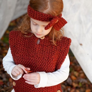 May include: A young girl wearing a red knitted vest with buttons and a matching red headband. The vest has a textured knit pattern and the headband is tied in a bow at the back.