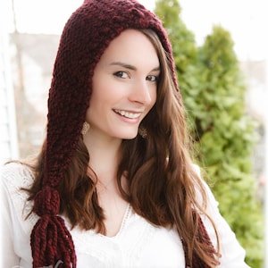 Peut inclure: Une femme portant un bonnet à capuche en tricot bordeaux avec de longues franges. Le bonnet est fait d'un fil doux et texturé et a une coupe ample et fluide.