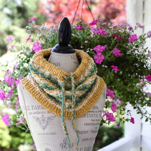 May include: A crocheted cowl with a striped pattern in shades of yellow, green, and white. The cowl is displayed on a mannequin with a decorative base.