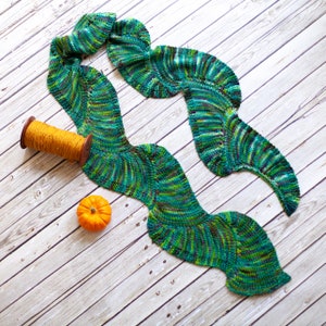 Può includere: Una sciarpa di lana verde e blu con un motivo ondulato. La sciarpa è appoggiata su una superficie di legno bianco con un gomitolo di lana e una piccola zucca arancione.