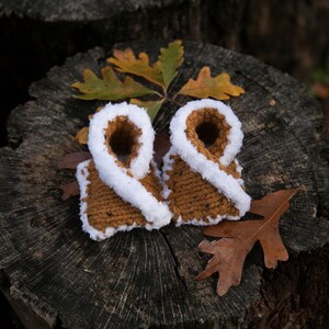 May include: A pair of brown knitted baby booties with white fur trim. The booties are sitting on a tree stump with fall leaves.