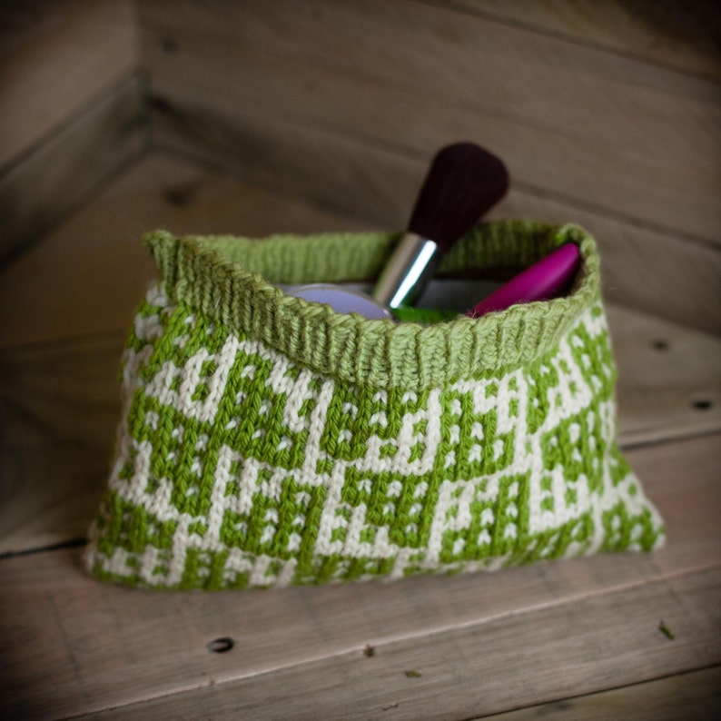 May include: A small, knitted bag in shades of green and white, with a geometric pattern. The bag's rim is a solid green, and it contains makeup brushes and other cosmetic items. The bag sits on a wooden surface.