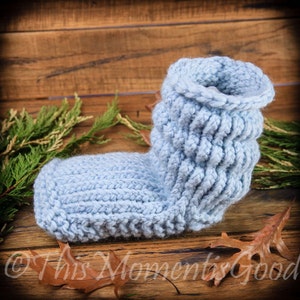 May include: A pair of light blue knitted slipper socks with a textured knit pattern. The socks are on a wooden surface with some leaves.
