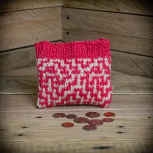 May include: A small, knitted pouch in shades of pink and white, with a geometric pattern. The pouch is sitting on a wooden surface, with several coins scattered in front of it. The pouch is likely used to store small items.