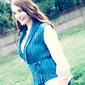 May include: A teal blue knitted vest with a shawl collar and a tie at the waist. The vest is worn over a white shirt.