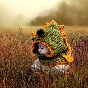 May include: A child wearing a green and orange crocheted dinosaur hat with a hood. The hat has a textured, knitted design and is worn over the child's head and neck.