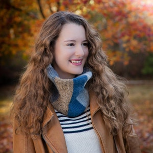 May include: A woman with long, curly brown hair is wearing a brown leather jacket, a striped sweater, and a blue and brown knit neck warmer. She is standing in front of a blurred background of fall foliage.