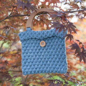 May include: A blue knitted tote bag with a wooden handle and a wooden button. The bag is hanging from a tree branch with red leaves.
