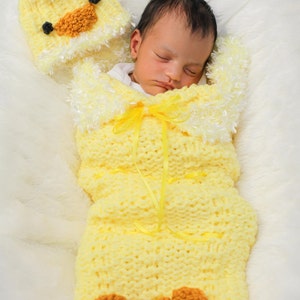 Puede incluir: Un bebé recién nacido duerme plácidamente en un saco de dormir de crochet amarillo con una capucha con forma de pato. El saco de dormir tiene una cinta amarilla atada alrededor del cuello y un ribete de piel blanca.
