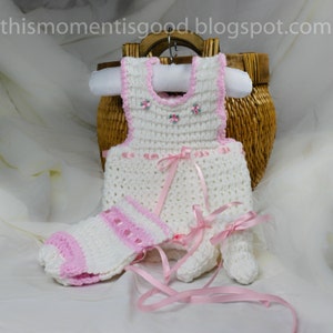 May include: White crocheted baby dress with pink ribbon accents and matching booties. The dress has embroidered flowers on the bodice.