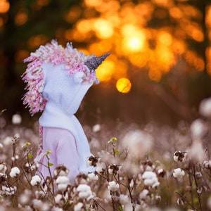 May include: A young person wearing a white hooded unicorn costume with pink and purple yarn hair stands in a field of white cotton bolls. The background is a blurred sunset.