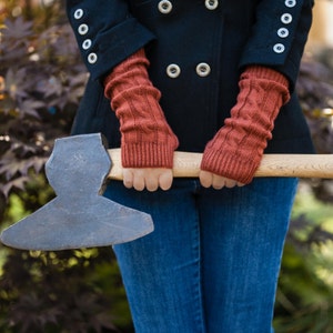 May include: A person wearing a black coat and blue jeans is holding a large, black axe with a wooden handle. The person is wearing red knitted arm warmers.