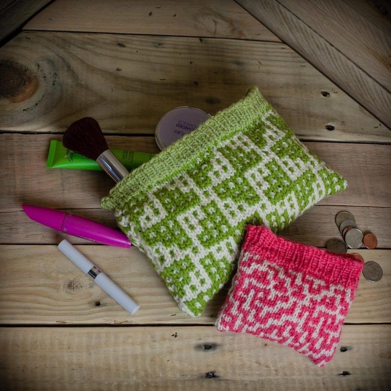 May include: Two knitted cosmetic bags, one green and white, the other pink and white, with geometric patterns. Makeup items, including a brush, mascara, and powder, are arranged around the bags on a wooden surface. A few coins are scattered nearby.