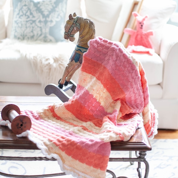 May include: A pink and white striped knitted baby blanket with a white crocheted border. The blanket is draped over a wooden rocking horse with a white and blue painted design.