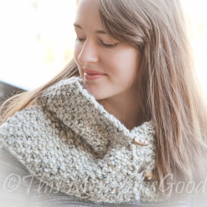 May include: A chunky knit cowl in shades of gray and cream, fastened with two wooden toggle buttons. The cowl is worn around the neck, offering warmth and a textured look. The text "This Art is Good" is visible in the bottom left corner.
