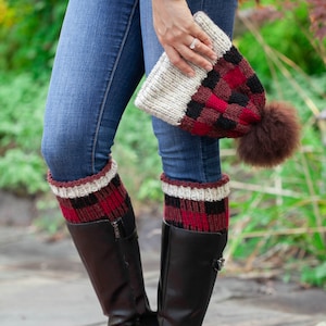 May include: A pair of black boots with red and black plaid knit leg warmers. The leg warmers have a white band at the top. The person is also wearing blue jeans and a red and black plaid knit hat with a white band and a brown fur pom-pom.