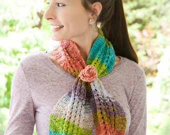 May include: A colorful knitted scarf with a flower accent. The scarf is made with a variety of colors, including green, blue, pink, and orange. The scarf is tied around the neck of a person wearing a purple shirt.