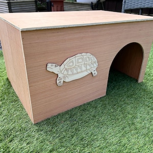May include: A wooden tortoise house with a cutout of a tortoise on the front. The house has a rounded opening on the side for the tortoise to enter.