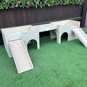 May include: A wooden rabbit hutch with two levels and two ramps. The hutch is painted a light wood colour and has a small cutout of a rabbit on the top level. The hutch is sitting on a green artificial grass surface.