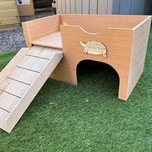 May include: A wooden pet habitat with a ramp and a raised platform. The habitat features a decorative wooden turtle on the side. The ramp has horizontal grooves for traction. The structure is made of light-coloured wood.