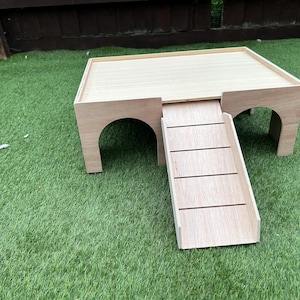 May include: A light-coloured wooden platform with two arched openings on the sides and a flat top surface. A detachable ramp with horizontal slats is positioned in the centre, connecting the ground level to the elevated platform surface.