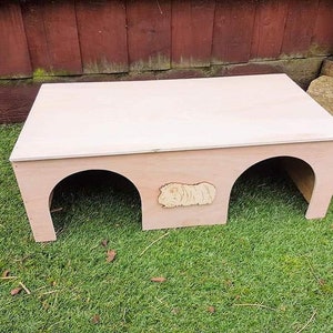 May include: A wooden pet house with two entrances and a cutout of a guinea pig on the front. The house is painted a light pink colour and is sitting on a green grassy surface.