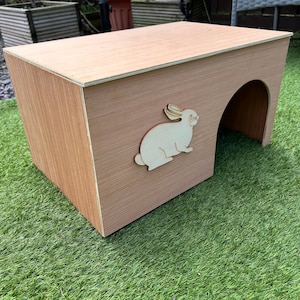 May include: A wooden rabbit hutch with a cutout of a rabbit on the front. The hutch has a rounded entrance and is made of natural wood.