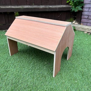 May include: A small, wooden house-shaped table with a light brown finish. The table has a sloped roof and arched openings on the sides. It is set on a green lawn.