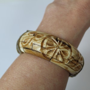 Op de afbeelding: Een gesneden houten armband met een bloemenontwerp. De armband is bruin met donkerbruine accenten en wordt om een pols gedragen. De armband is ongeveer 6,35 cm breed.