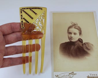 Puede incluir: Un peine para el cabello vintage de color ámbar con una parte superior decorativa adornada con pedrería oscura y un diseño ornamentado. El peine tiene cuatro púas largas. Un retrato en tonos sepia de una mujer está al lado del peine.