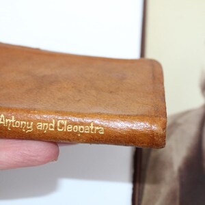Peut inclure: Un petit livre en cuir marron avec le titre "Antony and Cleopatra" en lettres dor&eacute;es. Le livre est tenu dans une main, avec un portrait en s&eacute;pia en arri&egrave;re-plan.