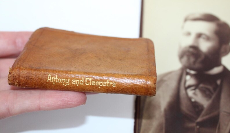 Peut inclure: Un petit livre en cuir marron avec le titre "Antony and Cleopatra" en lettres dor&eacute;es. Le livre est tenu dans une main, avec un portrait en s&eacute;pia en arri&egrave;re-plan.