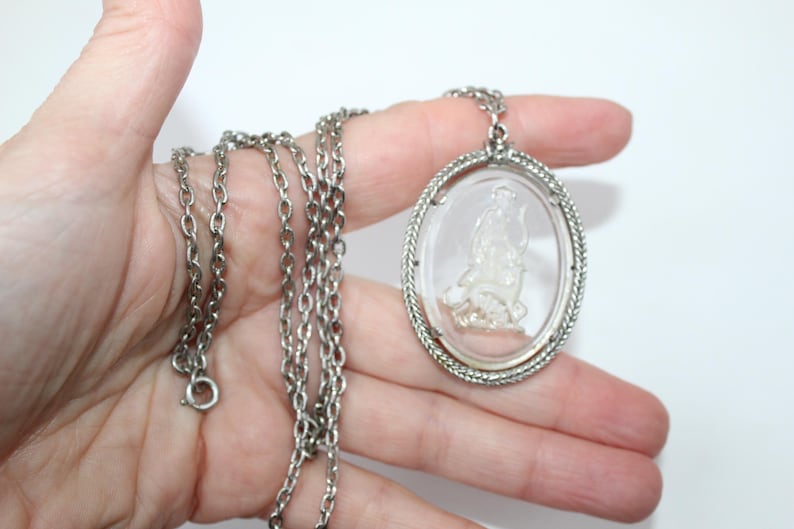 May include: Silver-toned necklace featuring an oval pendant with a clear, detailed cameo design. The pendant is framed with a textured border and hangs from a delicate chain. A classic piece of jewelry.