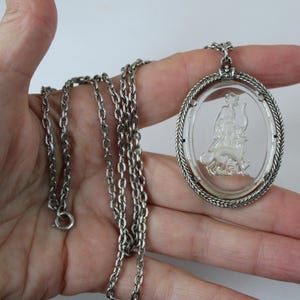 May include: Silver-toned necklace featuring an oval pendant with a clear, etched design of a figure riding an animal. The pendant is framed by a twisted rope-like border. The chain is a classic link style.