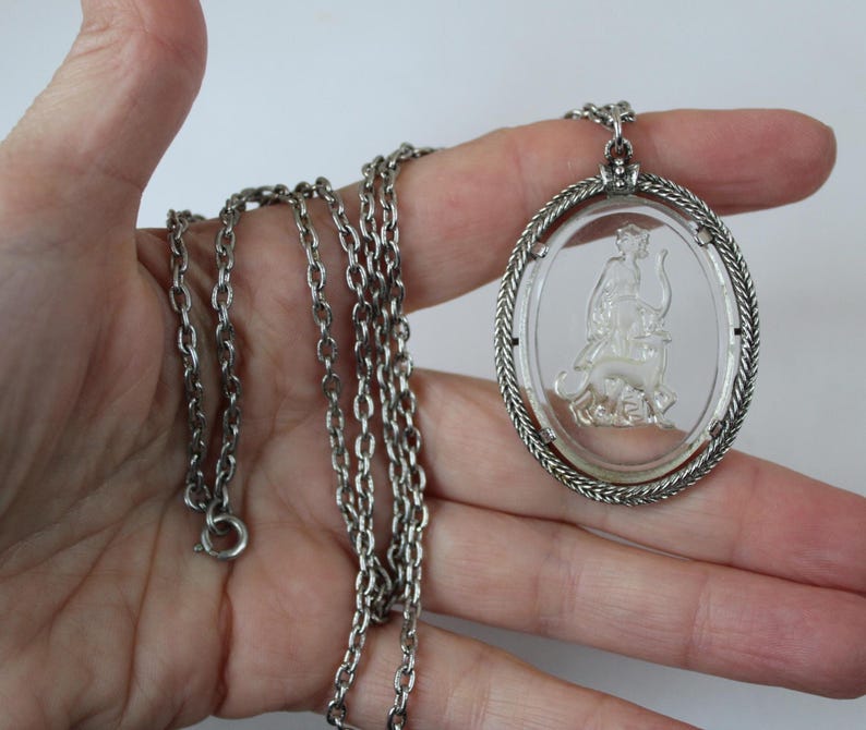 May include: Silver-toned necklace featuring an oval pendant with a clear, etched design of a figure riding an animal. The pendant is framed by a twisted rope-like border. The chain is a classic link style.