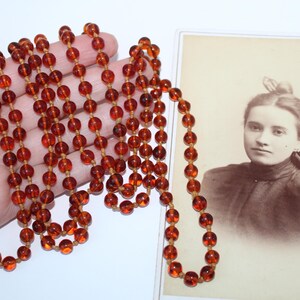 May include: A long necklace of amber-colored beads, held in a hand, next to a vintage portrait of a woman. The beads are round and strung on a light-colored cord. The portrait is in sepia tones.