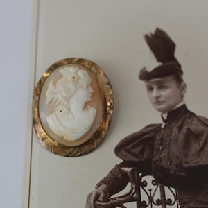 May include: Oval cameo brooch featuring a carved profile of a woman in cream and tan tones. The cameo is set in a gold-toned frame with an ornate, textured border. The background includes a sepia-toned photograph of a person.