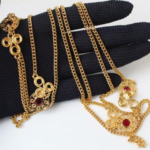 May include: A gold-tone metal chain necklace featuring multiple circular filigree links, each centered with a small, round, deep red gemstone. The necklace design incorporates intricate wire-work swirls and a classic curb chain link structure.