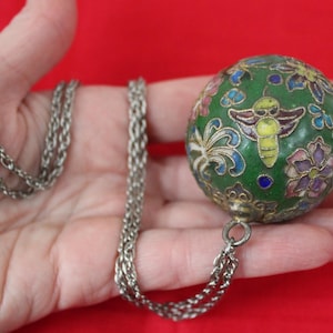 May include: A green, round pendant with a silver chain. The pendant features a bee and floral design in various colors. The chain is made of silver metal.