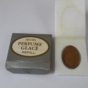 Pode incluir: Uma caixa de cartão cinzenta com a etiqueta AVON PERFUME GLACÉ REFILL com um centro oval castanho e um suporte de plástico branco contendo uma recarga de perfume oval castanha sólida. A caixa tem o texto 275-56-13 e 075040 na borda.