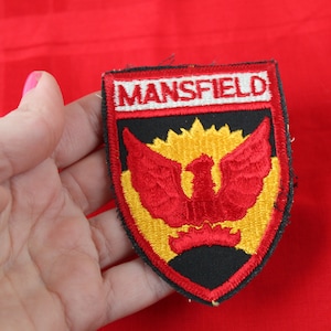 May include: A black, red, and yellow embroidered patch with the word "MANSFIELD" in white lettering. The patch features a red phoenix with yellow flames.