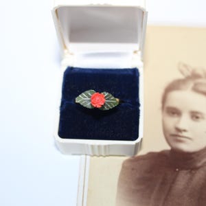 Può includere: Un anello con un piccolo bocciolo di rosa rossa centrato tra due foglie verdi, presentato all'interno di un portagioie bianco con rivestimento in velluto blu scuro. L'anello è posizionato accanto a una fotografia color seppia di una persona.