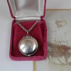 May include: A silver locket necklace with an engraved initial, presented in a pink velvet jewellery box. The locket hangs on a delicate chain. A vintage photograph is in the background, adding a nostalgic touch to the presentation.