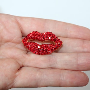Puede incluir: Broche de labios rojos de pedrería con borde dorado. Los labios están cubiertos de pequeños brillantes rojos brillantes. Un accesorio decorativo.