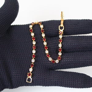 May include: A gold-tone tennis bracelet featuring an alternating pattern of round, faceted red and clear white stones set in a metal chain. The bracelet includes a secure metal clasp mechanism at one end and a small rectangular loop at the other.