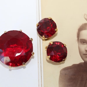 May include: A set of three vintage ruby red rhinestone jewelry pieces. The largest is a round brooch, and the other two are smaller, round earrings. All pieces have a gold-tone metal setting. A sepia-toned photograph of a woman is in the background.