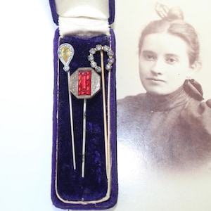May include: Three antique stick pins in a purple velvet case. The pins feature various gemstone designs, including a yellow teardrop, a red rectangular stone, and a circle of small clear stones. A vintage photograph of a woman is in the background.