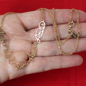 May include: A gold-toned necklace featuring multiple interconnected metal links, each shaped into the cursive word Amour. The chain includes a circular spring ring clasp and a small rectangular metal tag attached near the closure mechanism.